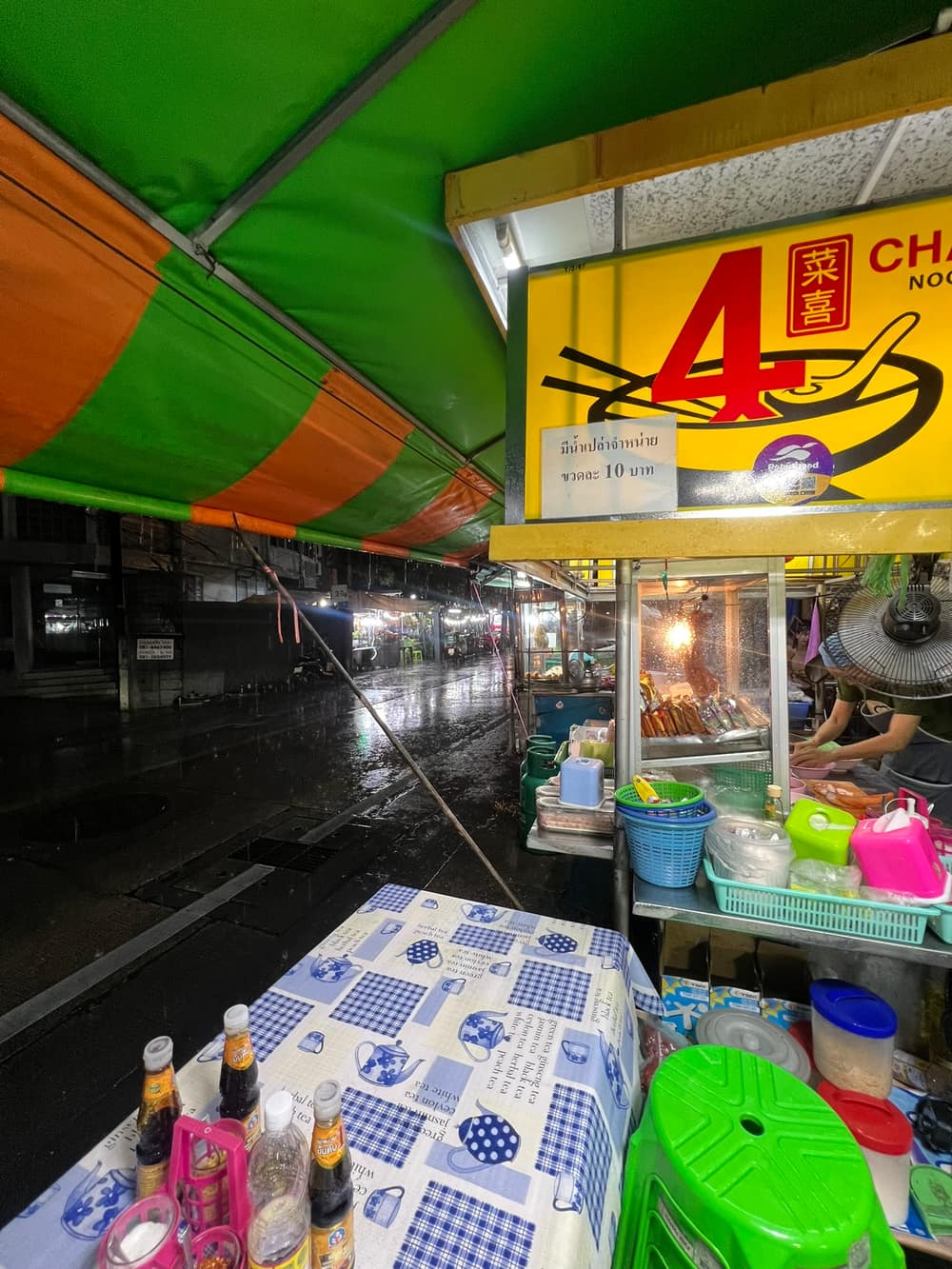 A cart in Soi Si Bamphen at night with a yellow Chaixi noodle sign, wet street and plastic stools