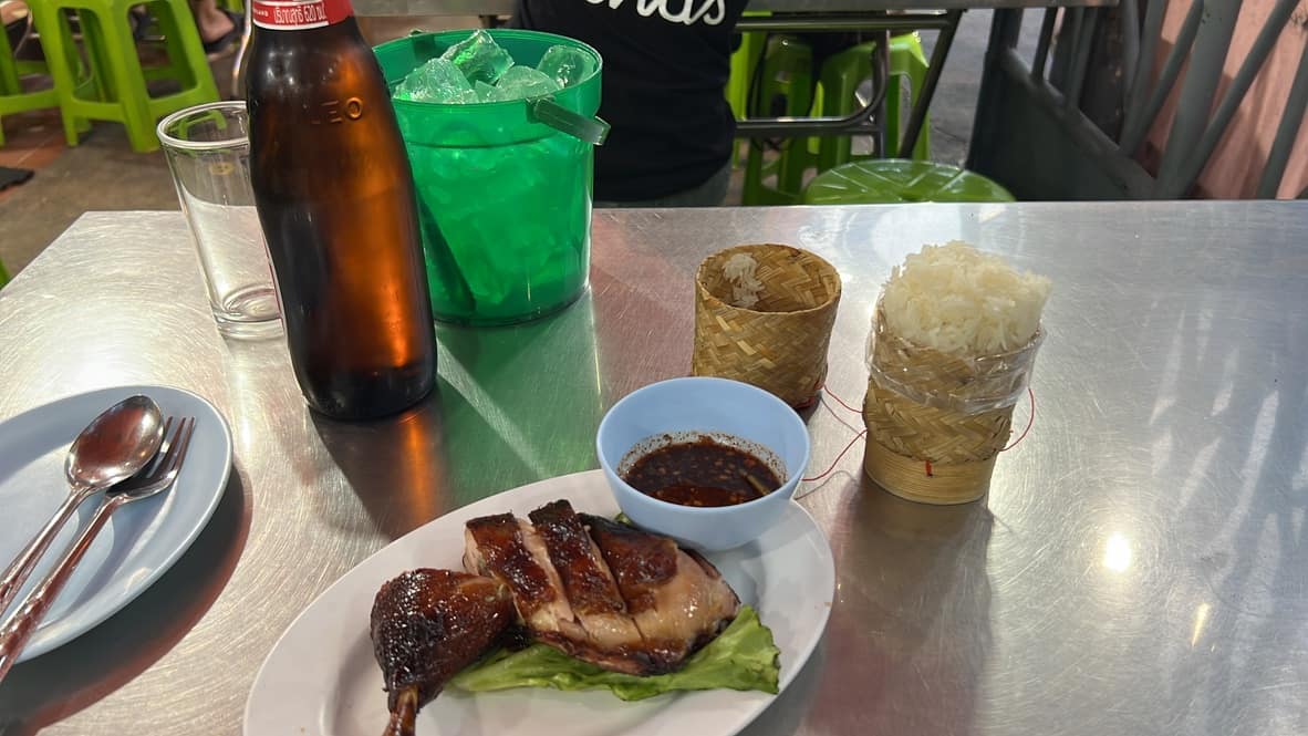 Grilled chicken, sticky rice in bamboo baskets, dipping sauce and a bottle of Leo beer on a steel table