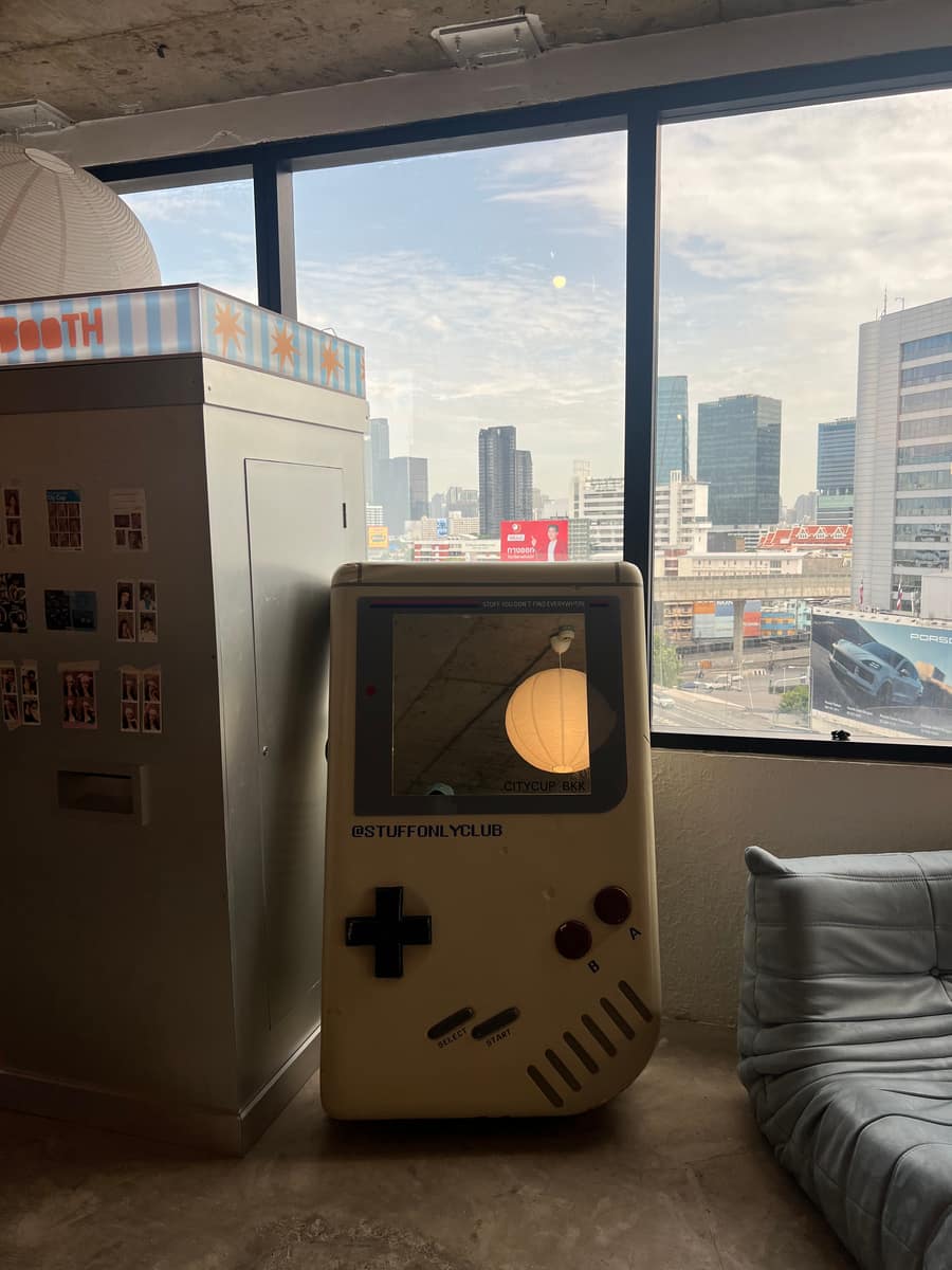 A giant Game Boy standing beside the photo booth at Citycup BKK, next to a window looking over the city