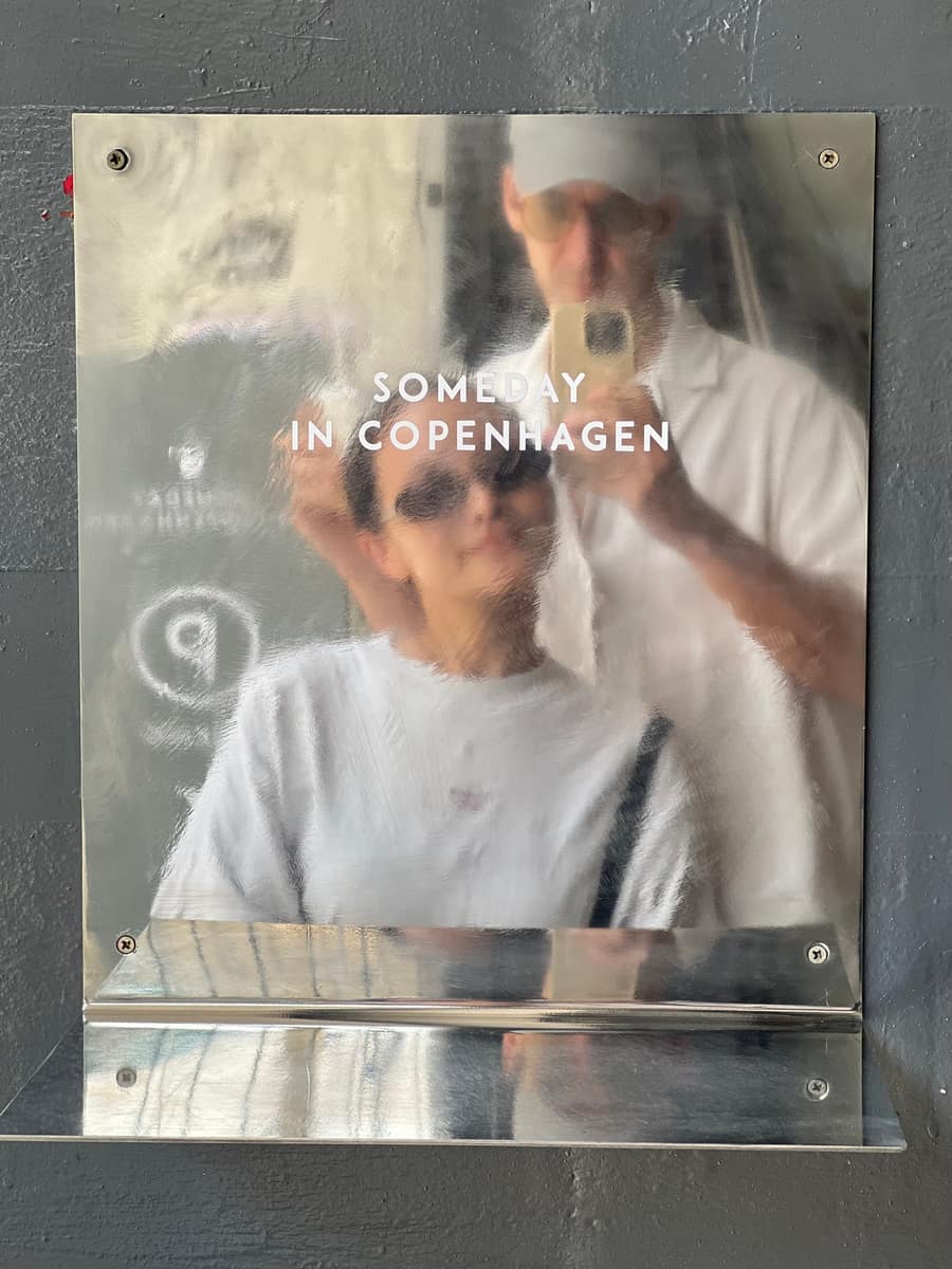 A polished-metal Someday in Copenhagen sign on a grey wall