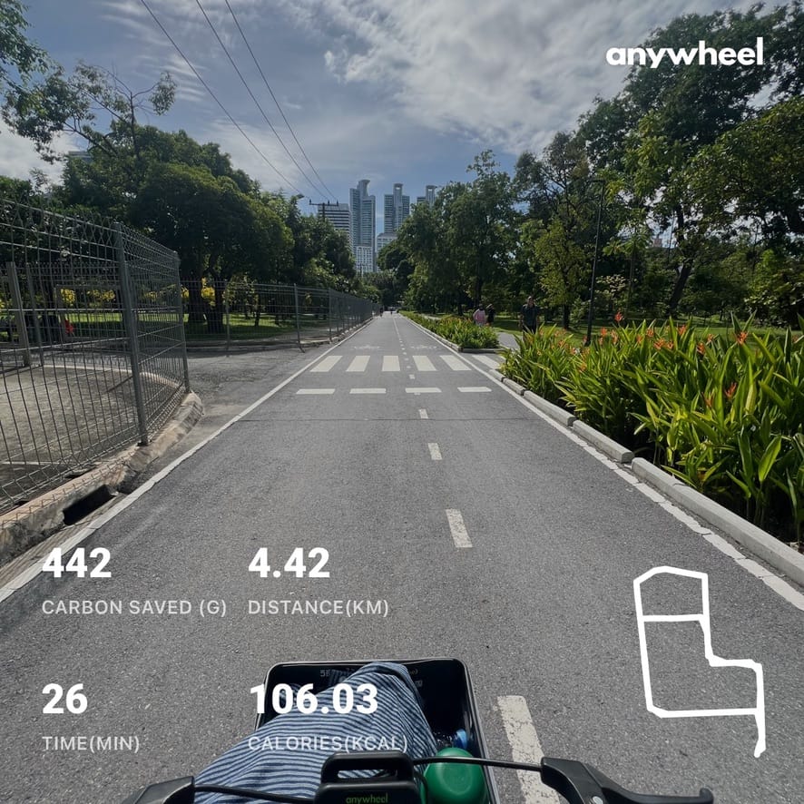 First-person view cycling a green Anywheel bike down a park path with the Bangkok skyline ahead